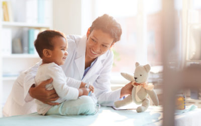 Young female doctor in white coat showing toy to her little patient and playing with him at hospital