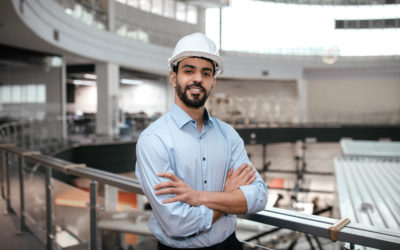 Cheerful confident millennial middle eastern male engineer in protective helmet with beard crossed arms on his chest controls work in office factory interior. Industry, business, covid-19 outbreak