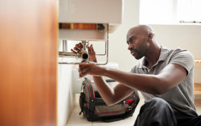 Young black male plumber sitting on the floor fixing a bathroom sink, seen from doorway